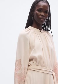 Lyserød bluse med lange ærmer og diskret blomstermønster på ærmerne. Blødt stof med plissert detalje og matchende bælte i taljen.