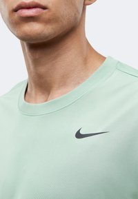 Κοντινό πλάνο ενός ατόμου που φοράει ένα ανοιχτοπράσινο T-shirt της Nike με μαύρο λογότυπο Swoosh στο στήθος, μπροστά σε λευκό φόντο.