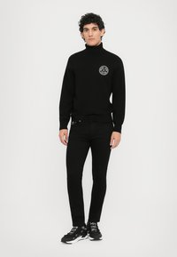 Μαύρη ζακέτα με ψηλό λαιμό και λογότυπο εμπρός, συνδυασμένη με μαύρα στενά τζιν και μαύρα αθλητικά παπούτσια με λευκές λεπτομέρειες και μοτίβο στα κορδόνια.