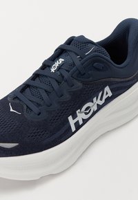 HOKA urheilusandaali, jossa on verkkokangas päällinen tummansinisessä värissä, paksu valkoinen välikerros ja sävy sävyyn nauhat, joissa on näkyvä logo ja teksturoituja yksityiskohtia.