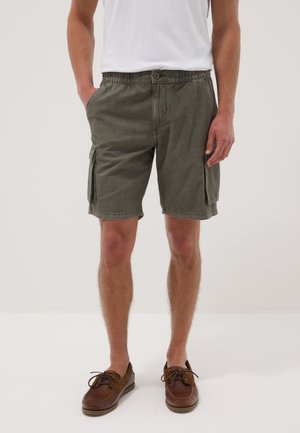 Mann trägt olivgrüne Cargo-Shorts mit Seitentaschen, weißes T-Shirt und braune Leder-Schnürschuhe, steht vor einfarbigem Hintergrund.