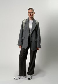 Blazer grigio in misto lana con design strutturato, abbinato a pantaloni marroni a vita alta e sneaker bianche. Zaino grigio chiaro sulle spalle.