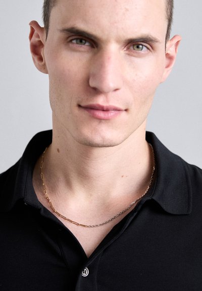 Αλυσίδα κολιέ με χρυσές και ασημένιες στρώσεις φορεμένη πάνω από ένα μαύρο πόλο μπλουζάκι με γιακά. Κοντινή λήψη που εστιάζει στην περιοχή του λαιμού και του στήθους.