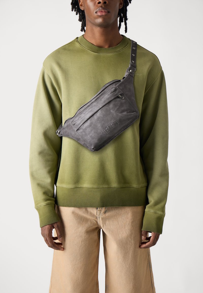 Grå lærcrossbody-veske med flere glidelåslommer, justerbar stropp og en strukturert overflate, båret over en olivengrønn hettegenser.