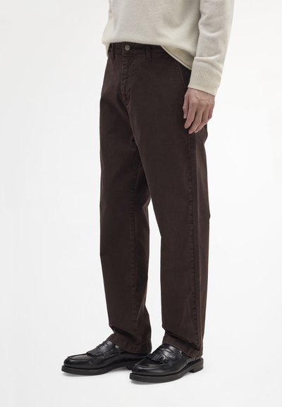 Pantalon en coton marron foncé avec une coupe droite, doté de poches avant et d'une fermeture par bouton. Associé à des mocassins noirs, affichant une texture lisse.