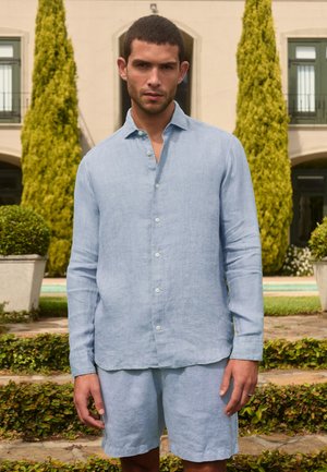 Uomo che indossa una camicia azzurro chiaro a maniche lunghe con bottoni e pantaloncini abbinati in piedi all'aperto davanti a un giardino curato e a un edificio.