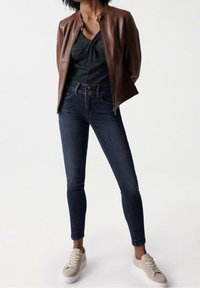 Braune Leder-Jacke mit Reißverschluss, grünem strukturiertem Oberteil, dunklen Skinny Jeans und beigen Sneakers. Fokus auf lässige, moderne Designelemente.