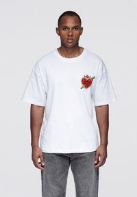 Weißes Baumwoll-T-Shirt mit kurzen Ärmeln, das ein rotes und goldenes, besticktes Herzmotiv auf der linken Brustseite zeigt. Lockerer Schnitt.