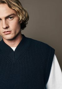 Marineblauwe gebreide vest met een V-halsontwerp en brede armsgaten, voorzien van een geribbelde textuur en gedragen over een wit shirt.