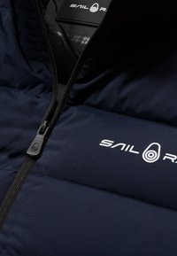 Mörkblå pufferjacka med svart dragkedja, vadderad design och en vit "SAIL R"-logotyp på bröstet för identifiering och branding.
