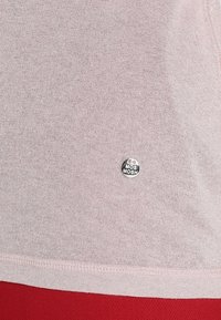 Blødt pink stof med en glat tekstur, der har et rundt sølv MOS MOSH logo som accent nær kanten, omkranset af rødt materiale nedenfor.