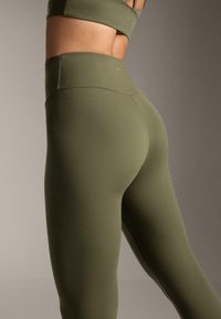 Közeli kép egy személyről, aki olívazöld, magasított derekú leggingset és hozzáillő sportmelltartót visel, hátulról és oldalról, semleges háttér előtt.