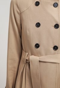Trench beige con botones negros de doble botonadura, cinturón tipo corbata y una textura de tela suave; presenta un corte ajustado y un diseño estructurado.