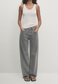 Débardeur blanc à bretelles fines, associé à un jean en denim gris à coupe ample et taille haute. Sandales noires sous des jambes de pantalon larges.