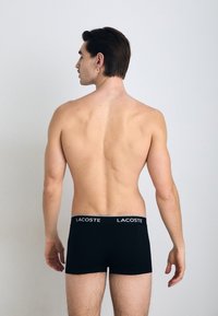 Boxers noirs avec le logo "LACOSTE" blanc sur la ceinture, portés par un mannequin masculin avec des cheveux courts, vus de dos sur un fond neutre.
