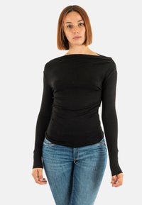 Giovane donna con capelli rossi lisci, che indossa una maglietta nera a maniche lunghe e jeans blu, in piedi di fronte a uno sfondo bianco semplice.