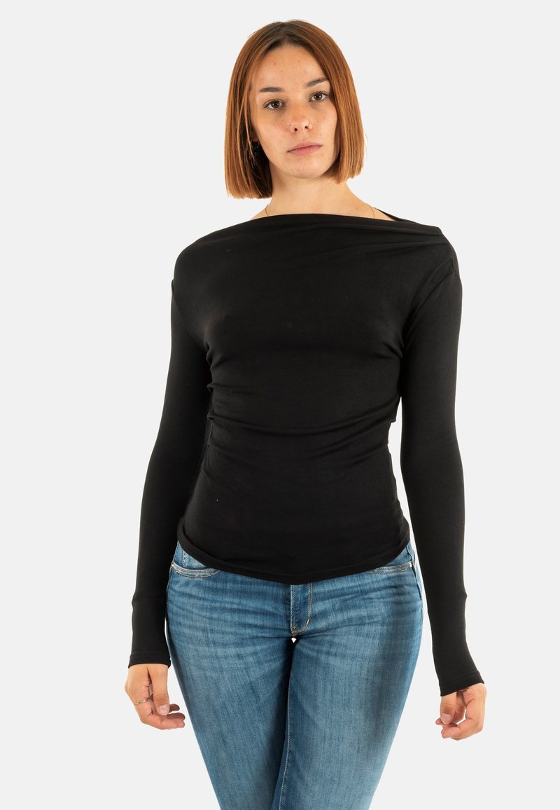 Giovane donna con capelli rossi lisci, che indossa una maglietta nera a maniche lunghe e jeans blu, in piedi di fronte a uno sfondo bianco semplice.