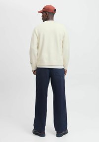 Man wearing cream sweater, navy wide-leg pants, black shoes, and red cap, standing facing away against plain white background.