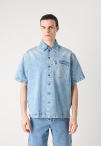 Camisa de denim de manga corta azul claro con cierre de botones en la parte delantera, bolsillo en el pecho y un ajuste relajado, con una textura desgastada y botones oscuros.
