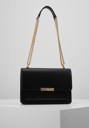 Czarna skórzana torebka z złotym łańcuszkiem i prostokątną złotą tabliczką z logo na przedniej klapie, wystawiona na białym piedestale na szarym tle.