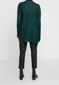 Długi, ciemnozielony kardigan o teksturowanym wykończeniu, w luźnym fasonie z okrągłym dołem, zestawiony z czarnymi tapered skórzanymi spodniami i botkami za kostkę.