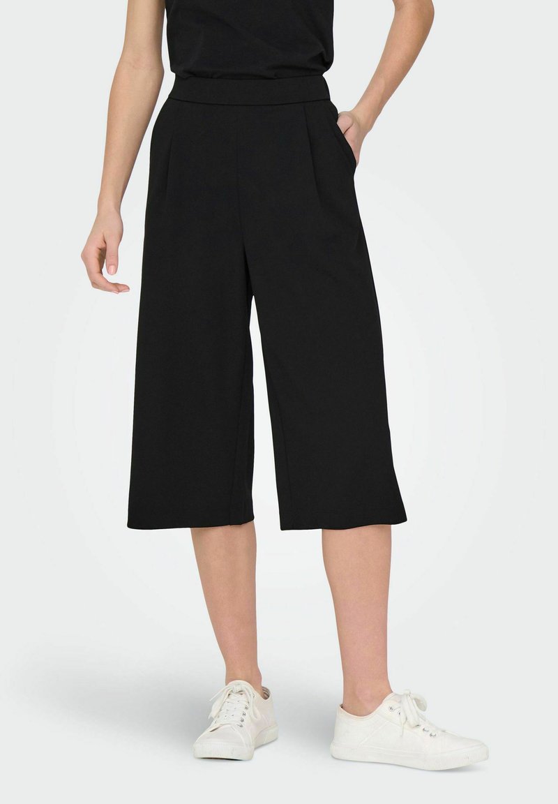 Svarta culotter med breda ben, hög midja och framvikt, gjorda av ett slätt tyg, i kombination med vita sneakers.