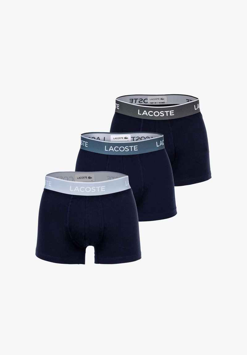 Tre paia di boxer Lacoste blu scuro con fascia elastica in grigio, blu e azzurro, ciascuno con il logo Lacoste.