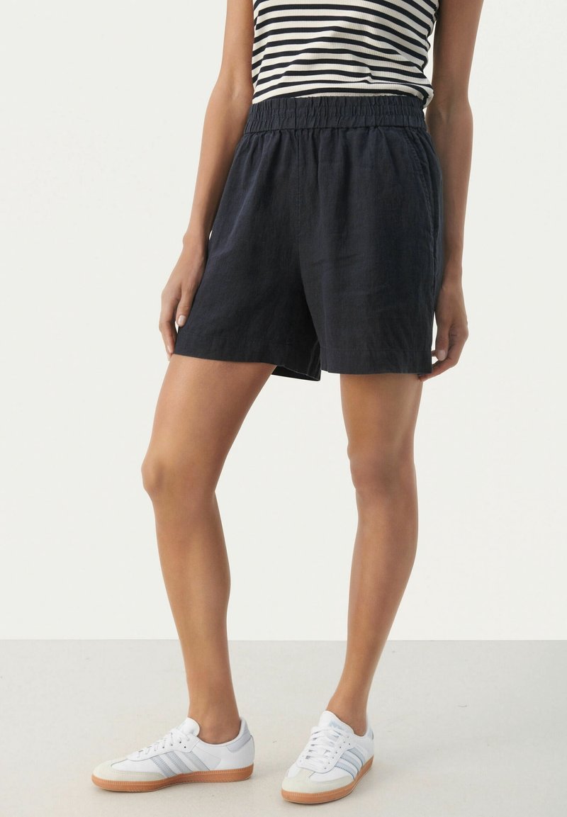 Shorts en lin bleu marine avec une taille élastique, présentant une coupe décontractée et des poches latérales. Associés à un débardeur rayé noir et blanc et des baskets blanches.
