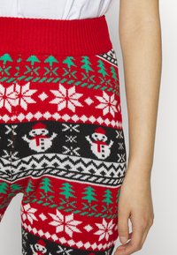 Leggings de malha com tema festivo, apresentando uma cinta canelada vermelha, com motivos de inverno em preto, branco e verde, incluindo flocos de neve e bonecos de neve.