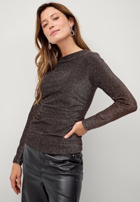 Maglia a maniche lunghe in tessuto marrone lucido, con una forma aderente e dettaglio arricciato sul lato. Abbinata a pantaloni in pelle nera.