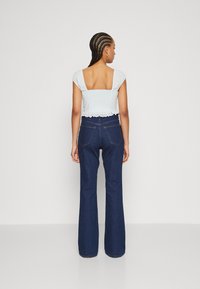 Top corto bianco con schiena quadrata e bordo in pizzo, abbinato a jeans blu scuro a zampa. Il tessuto ha una finitura testurizzata.