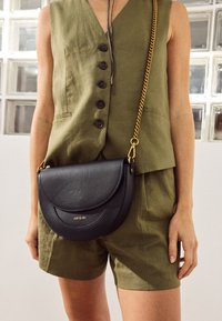 Borsa a tracolla in pelle nera dalla forma semicircolare, superficie testurizzata, hardware dorato e logo sul davanti; indossata con un outfit di colore verde oliva.