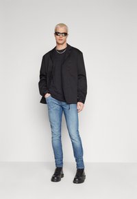 Jeune homme aux cheveux blonds courts portant des lunettes de soleil noires, une veste noire, un t-shirt, un jean bleu et des bottes noires, se tenant contre un fond blanc.