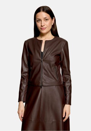 Femme aux longs cheveux foncés portant une veste zippée en cuir marron foncé et une jupe assortie, faisant face à l'avant avec une expression neutre.