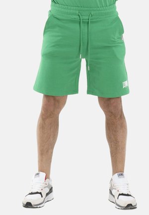 Homme portant un short vert vif à cordon de serrage et des baskets blanches et noires, debout les jambes légèrement écartées sur un fond uni.