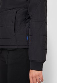 Svart kviltad jacka med ett diamantmönster, sidoficka, ribbad mudd och en blå detalj nära fållen. Slät textur.