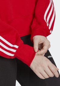 Red sweatshirt with white three-stripe design on sleeves. Ribbed cuffs and hem, soft texture. Worn with black pants featuring logo detail.