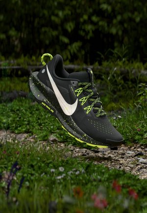 Schwarzer und neongrüner Nike Trail-Laufschuh mit weißem Swoosh, dargestellt über einem Kiesweg, umgeben von Gras und Wildblumen.