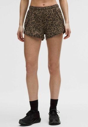Frau trägt leopardgemusterte Sportshorts, schwarze Knöchelsocken und schwarze Laufschuhe, steht vor einem schlichten Hintergrund.