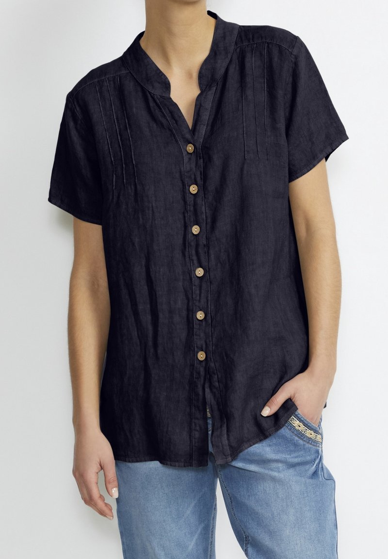 Schwarz, kurzärmlig, Leinenhemd mit Stehkragen, vertikalen Nähten und Holzknöpfen, kombiniert mit hellblauen Jeans.