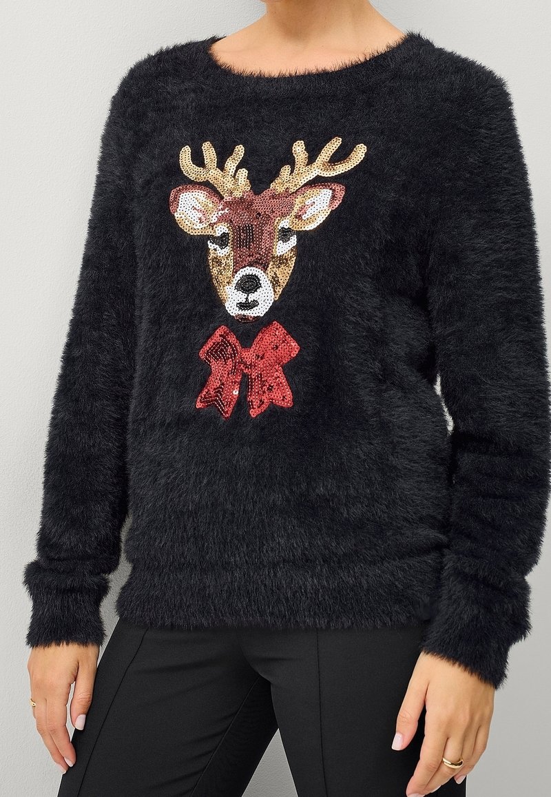 Pull noir à poils avec une tête de renne ornée de sequins et un nœud rouge, porté par une personne vêtue d'un pantalon noir et de bagues aux doigts.