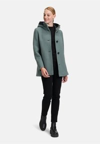 Grøn uldblandet jakke med hætte, to store sorte knapper, sidelommer og en blød tekstur, parret med sorte bukser og kraftige støvler.