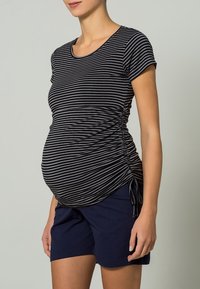 Czarna i szara w paski bluzka ciążowa z krótkimi rękawami, z bocznymi marszczeniami dla lepszej regulacji, w połączeniu z granatowymi szortami. Miękka tekstura materiału.