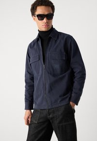 Mornarsko plava zip-up jakna s dva prednja džepa i logom. Nosiva preko crnog rolka-golfića, u kombinaciji s tamnim trapericama.
