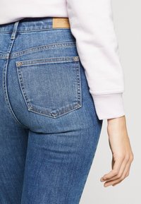 Jeans de denim azul con una forma ajustada, diseño de cinco bolsillos y etiqueta de cuero beige en la cintura trasera. La textura es suave con un ligero desgastado.