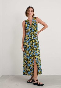 Rochie maxi florală cu un model de frunze albastre și galbene, design fără mâneci, decolteu adânc în V și o fante în față. Poartă sandale negre cu barete.
