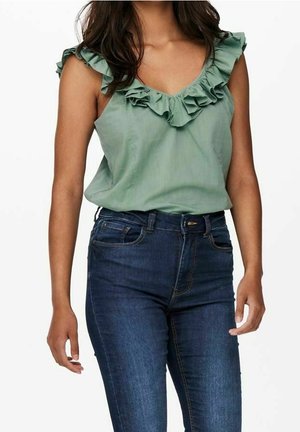 Nainen pukeutuneena hihattomaan salvianvihreään röyhelöiseen puseroon, joka on tungettu tummansinisiin korkeavyötäröisiin kapaisiin farkkuihin.