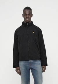 Black zip-up jacket with a high collar, textured finish, and a small yellow logo on the chest. Worn over blue denim jeans.