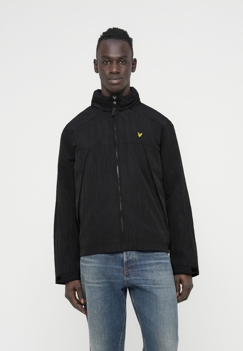 Black zip-up jacket with a high collar, textured finish, and a small yellow logo on the chest. Worn over blue denim jeans.