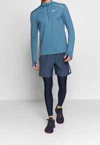 Niebieska sportowa bluzka z długim rękawem i zamkiem przy kołnierzu, w połączeniu z ciemnymi szortami i granatowymi legginsami. Czarnie sneakersy z fioletowymi akcentami dopełniają wygląd.